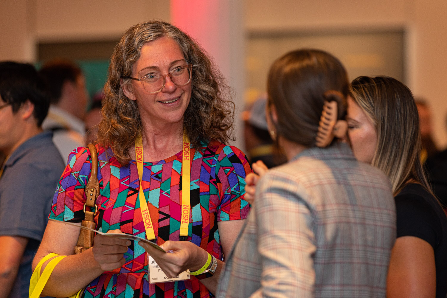 Woman with curly hair and glasses in a colorful geometric dress and yellow lanyard holding a card among a crowd.