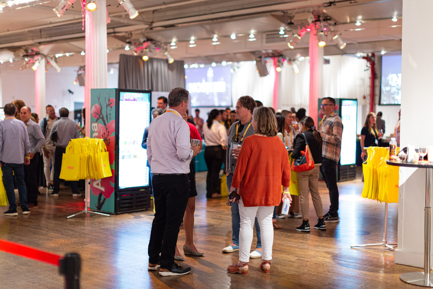 Wide room view of people standing and talking under pink-lit columns, Arizona coolers and yellow tote bags in the background.