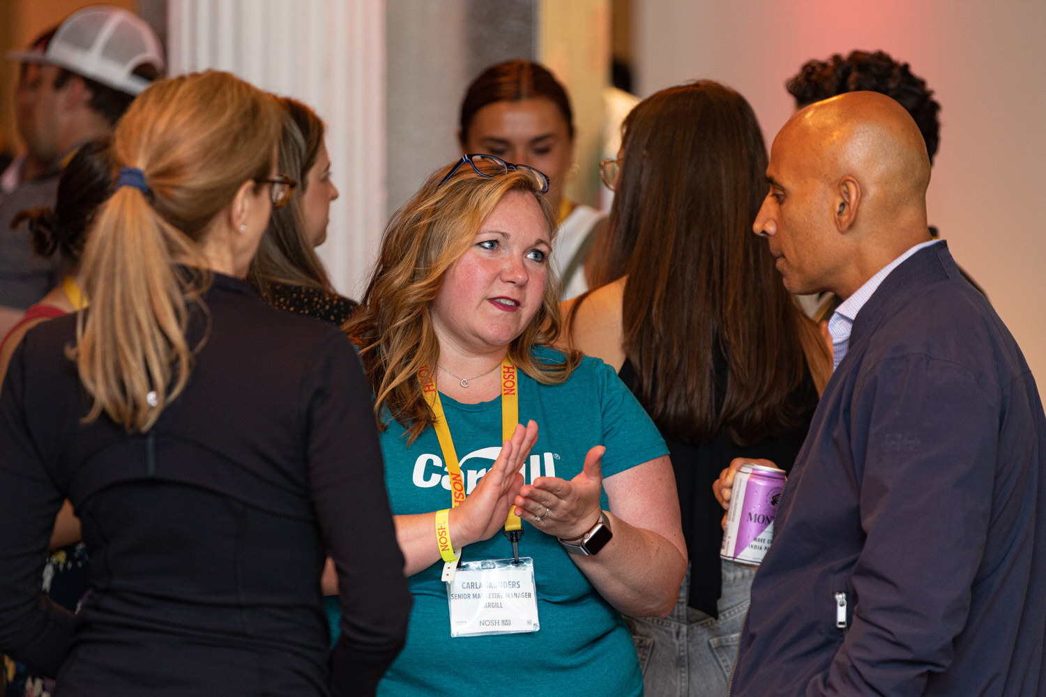 Woman in a teal Cargill shirt with a lanyard talking with a man in a dark jacket holding a can, others around them.