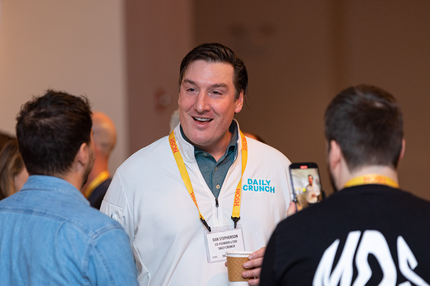 Man in a white Daily Crunch pullover with a lanyard talking with two men, one in a black MDS shirt.