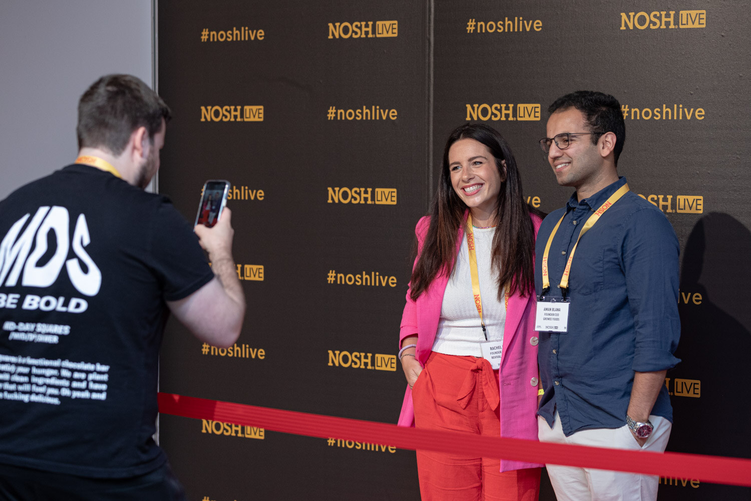 Woman in a pink blazer and man in a blue shirt in front of the #noshlive wall, a man in a black MDS shirt photographing them.