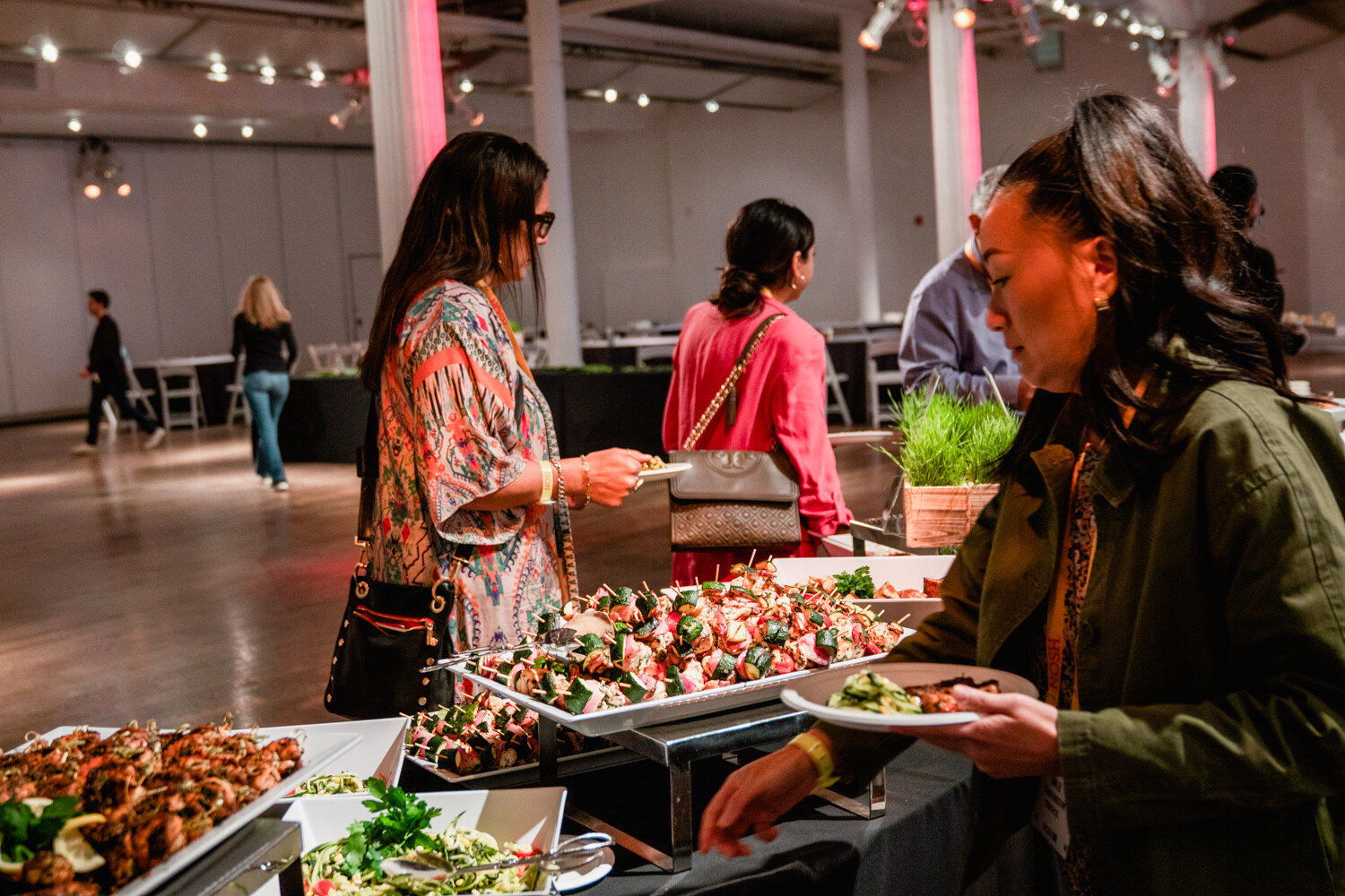 Woman in an olive jacket filling a plate beside trays of grilled chicken and salad while others wait.