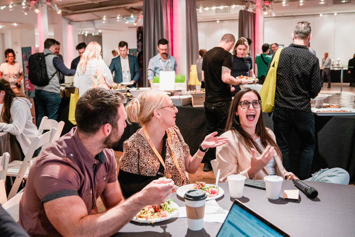 Woman in a leopard-print top and woman in glasses laughing at a table with plates and coffee cups, a man leaning in beside them.