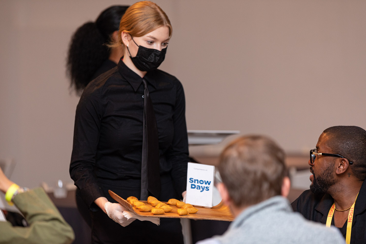 Server in a black mask holding a wooden tray of golden bites beside a Snow Days sign.