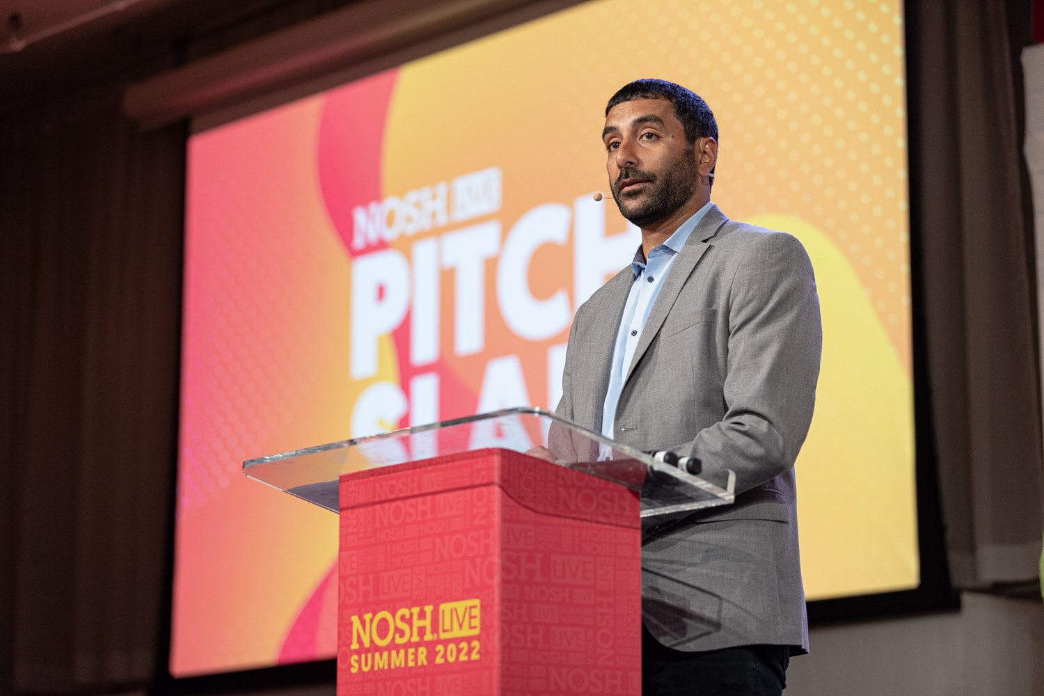 Man in a gray blazer speaking at a red Nosh Live Summer 2022 podium in front of an orange Pitch Slam screen.