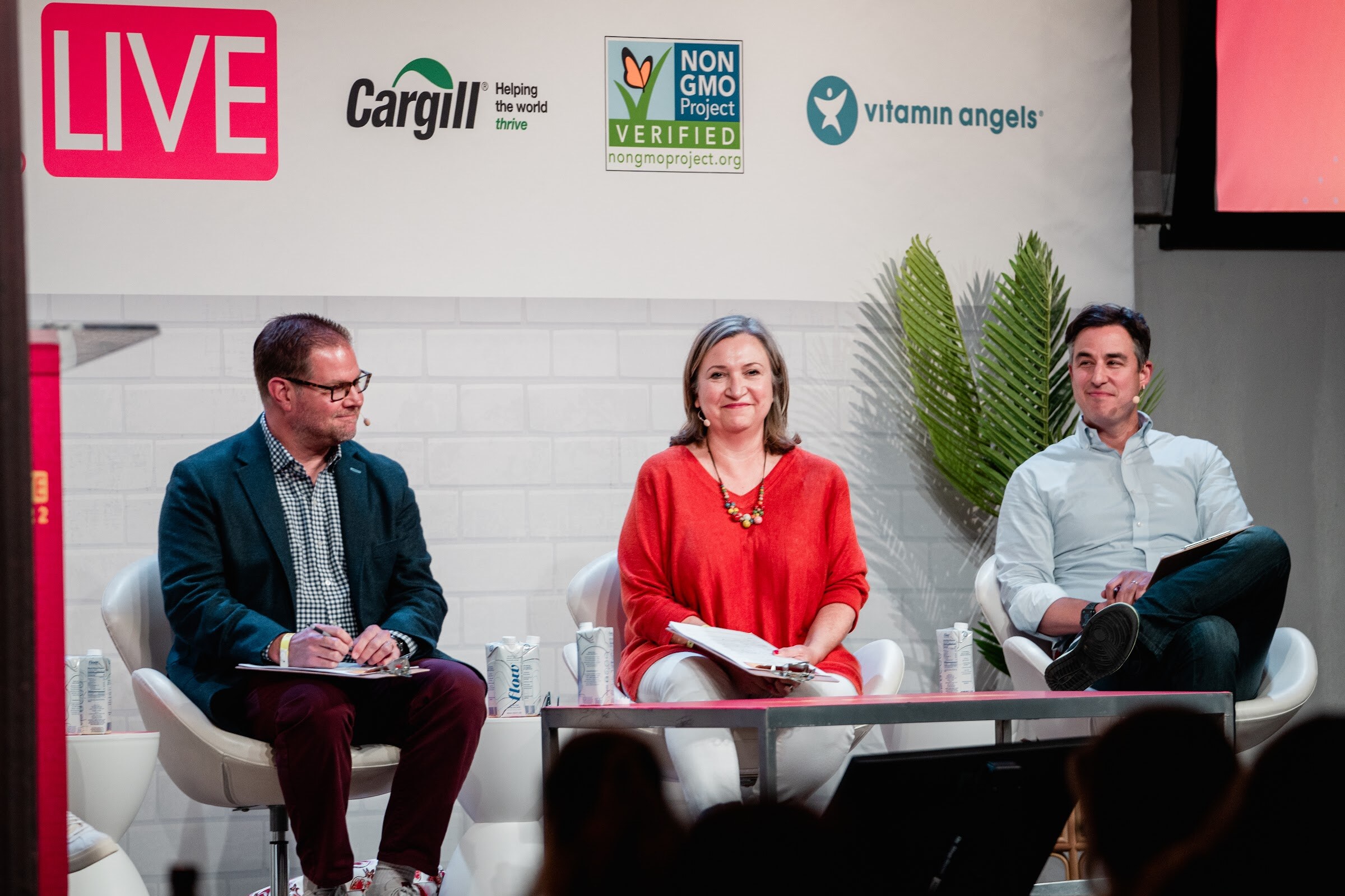 Man in a navy blazer, woman in a red top and man in a light shirt seated with notes under the Nosh Live and Cargill signs.