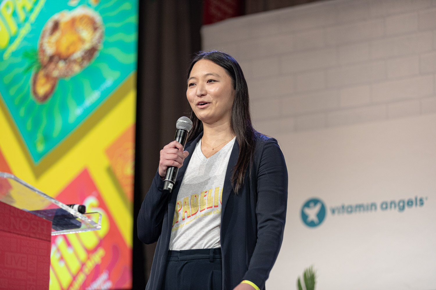 Woman in a dark blazer and a graphic tee holding a mic, with a colorful snack slide behind her.