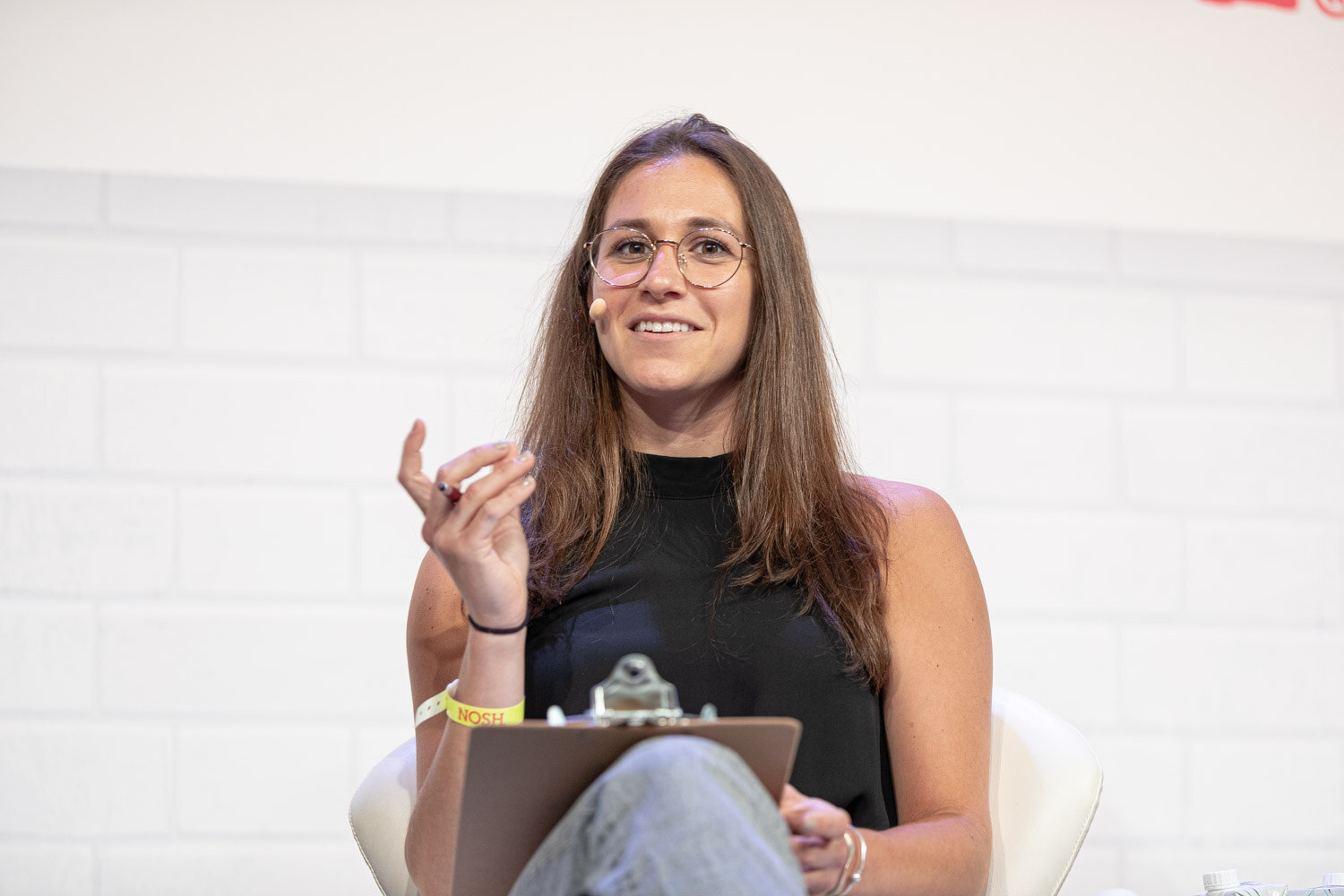 Woman with long brown hair and glasses in a black top, smiling with a clipboard on her lap.