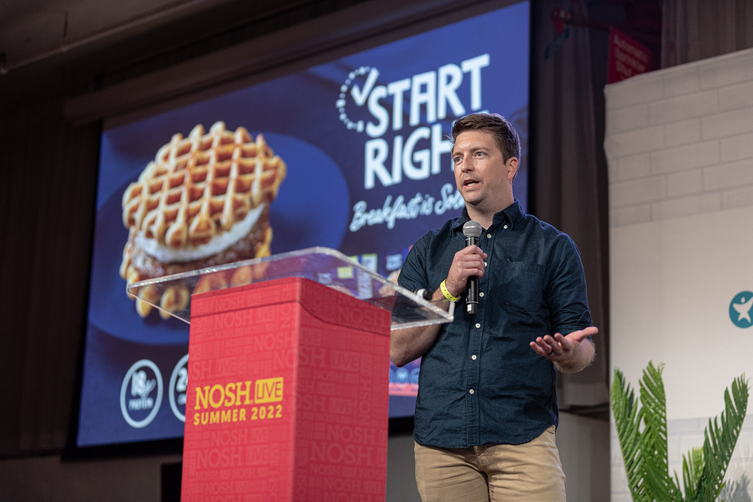 Man in a navy shirt with a mic beside a red podium in front of a Start Right waffle sandwich slide.
