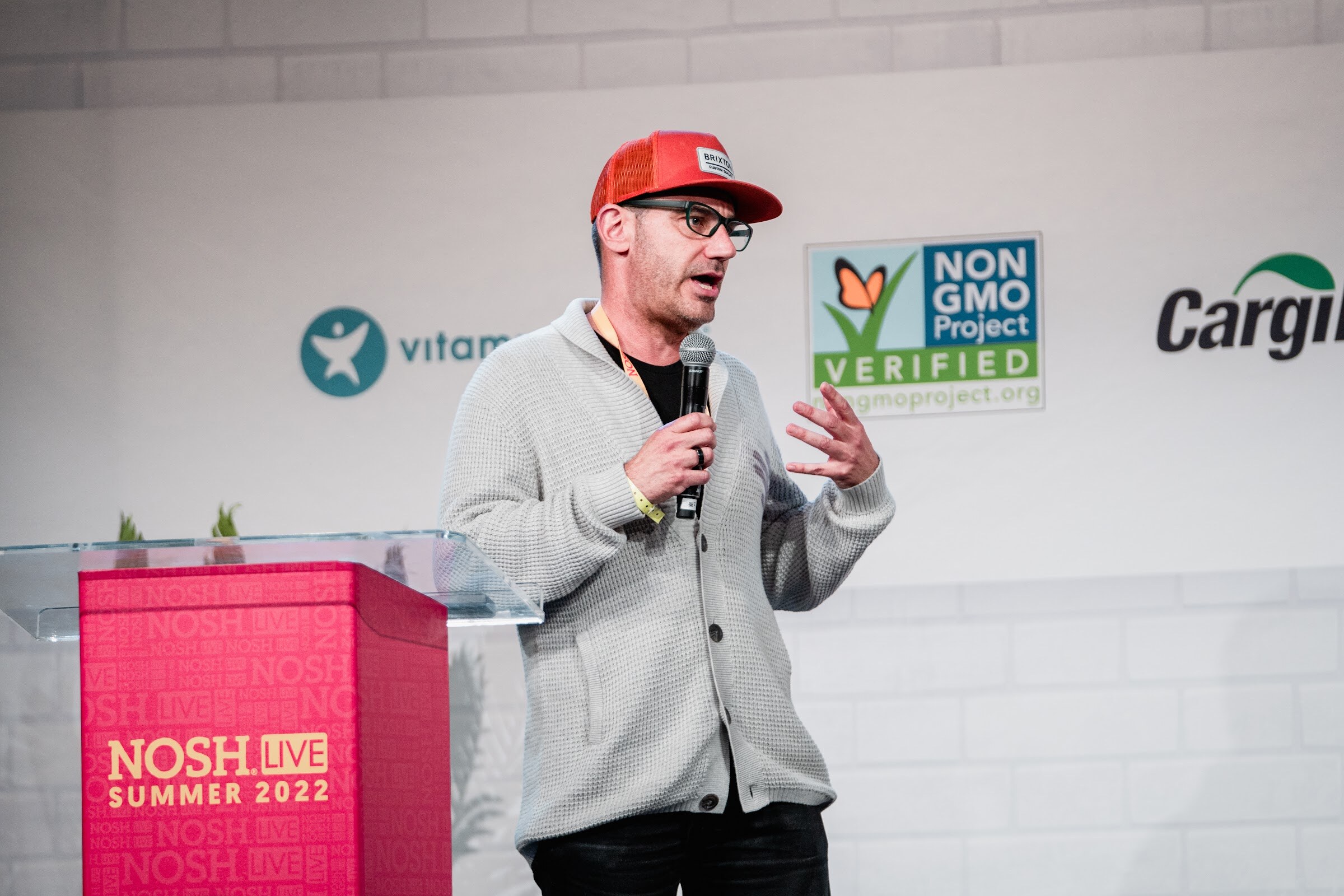 Man in an orange cap, glasses and a gray cardigan with a mic in front of a red podium and Non-GMO Project sign.