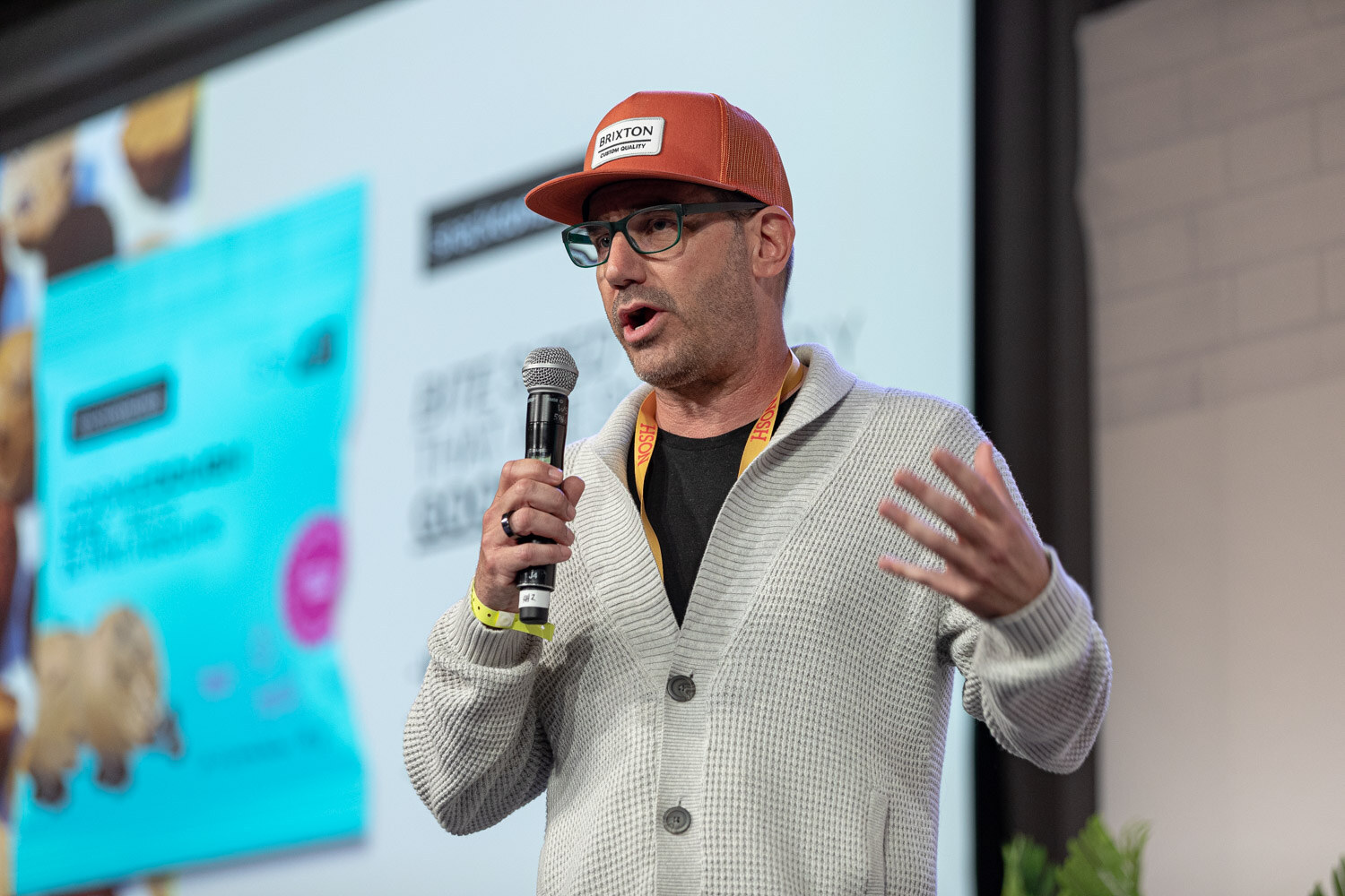 Man in an orange cap, glasses and a gray cardigan holding a mic with both hands raised, a snack slide behind him.
