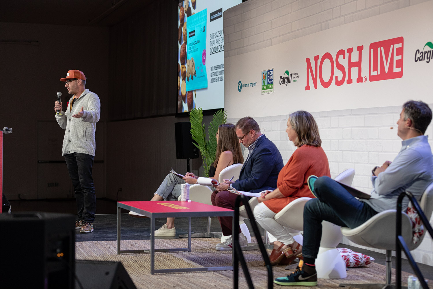 Man in an orange cap standing at left with a mic and three seated judges in front of a Nosh Live wall.