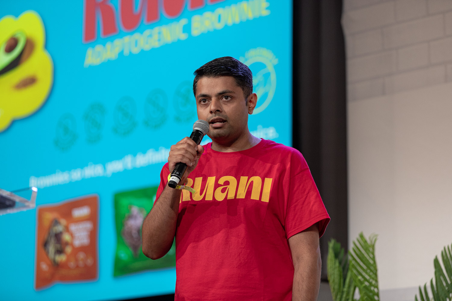 Man in a red Ruani t-shirt holding a mic in front of a bright blue slide reading Ruani Adaptogenic Brownie.