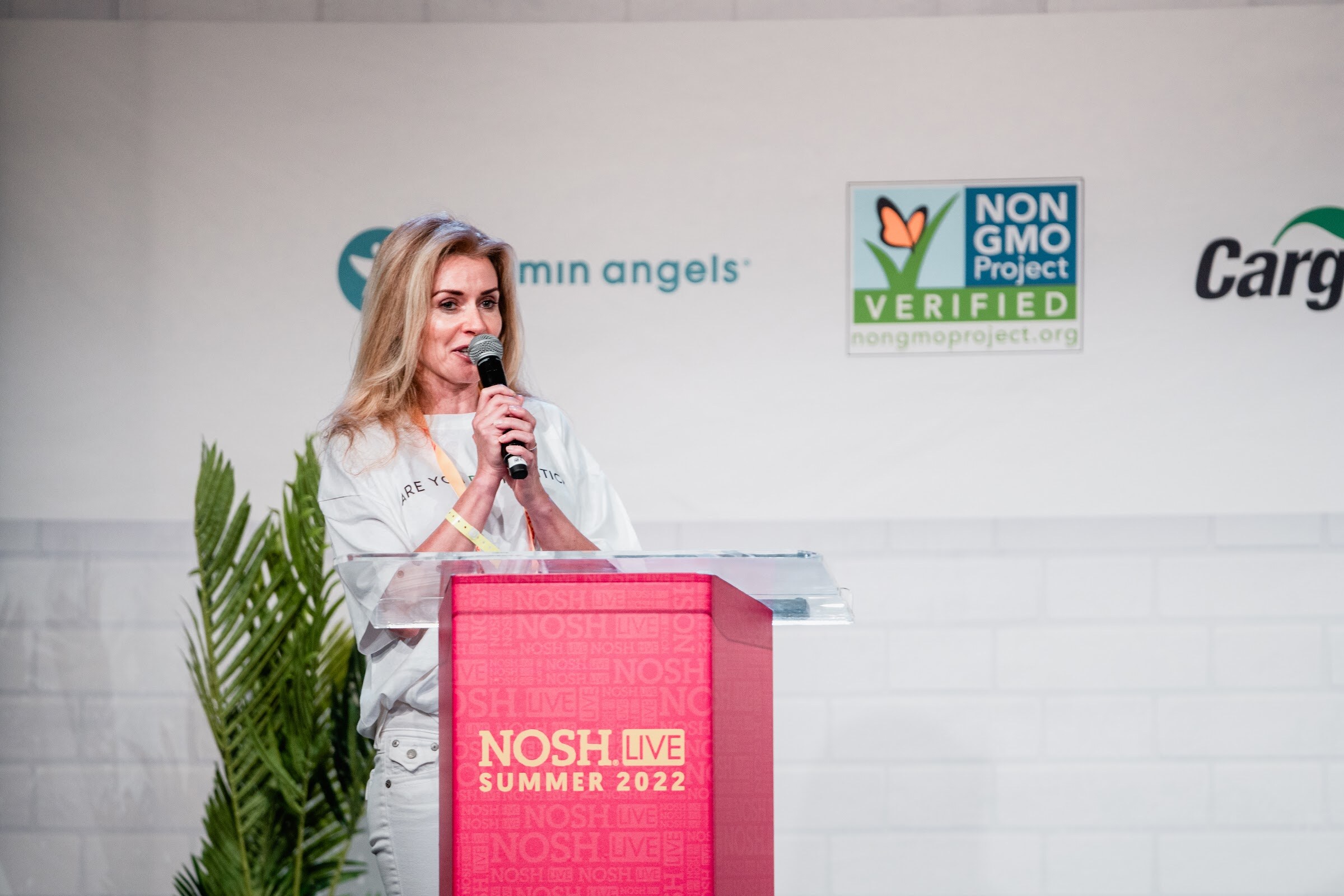 Blond woman in a white tee holding a mic at a red Nosh Live Summer 2022 podium.