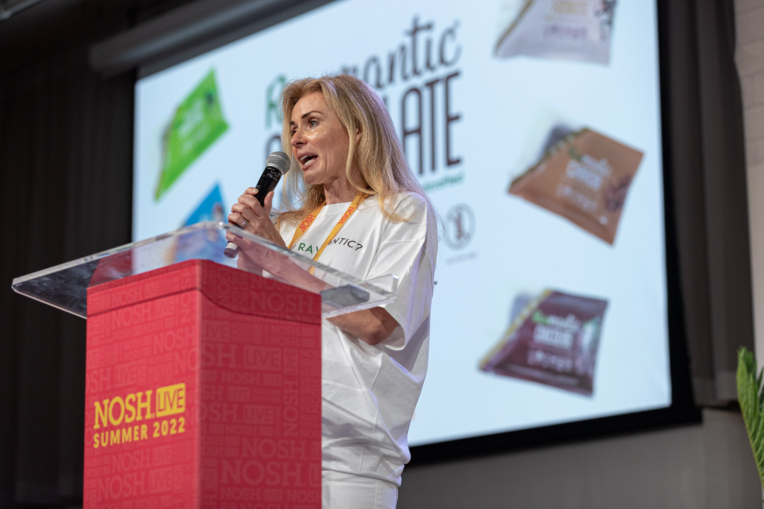 Blond woman in a white t-shirt and orange lanyard speaking into a mic at a red podium in front of a Rawmantic Chocolate slide.