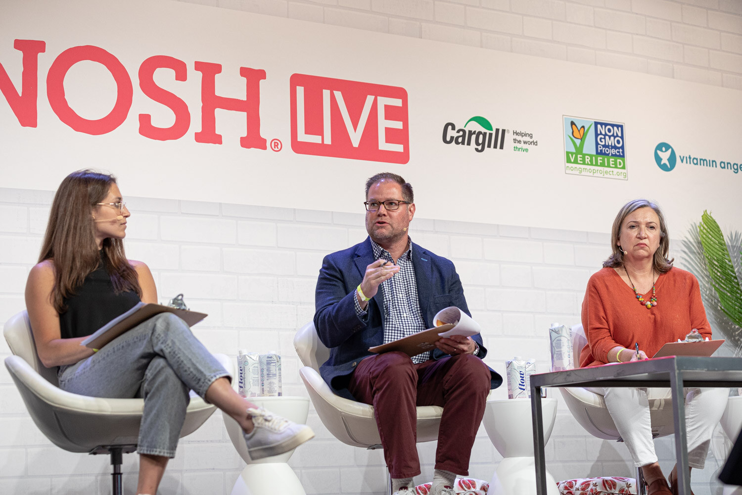 Woman in a black top, man in a navy blazer and woman in an orange top seated on stage in front of the Nosh Live wall.