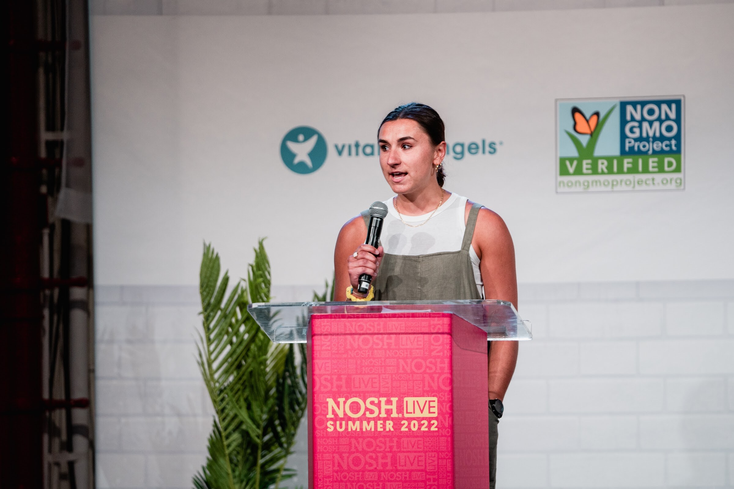 Dark-haired woman in olive overalls holding a mic at a red podium in front of a plant and a Non-GMO Project sign.