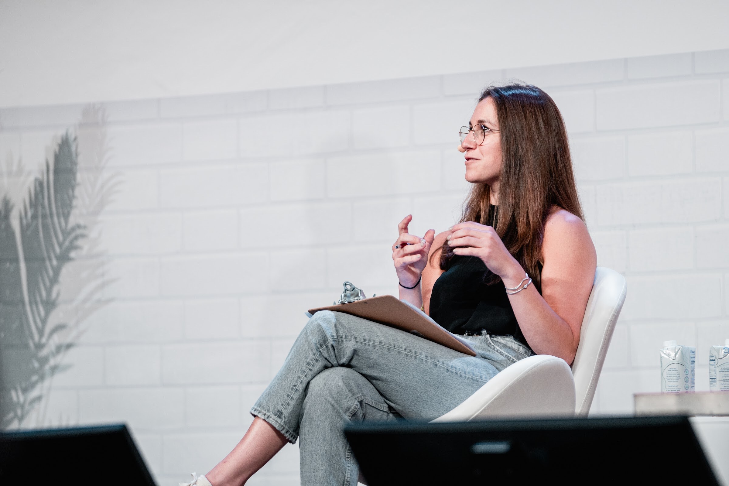 Woman with long brown hair and glasses seated on a white chair, hands up as she speaks, clipboard in her lap.