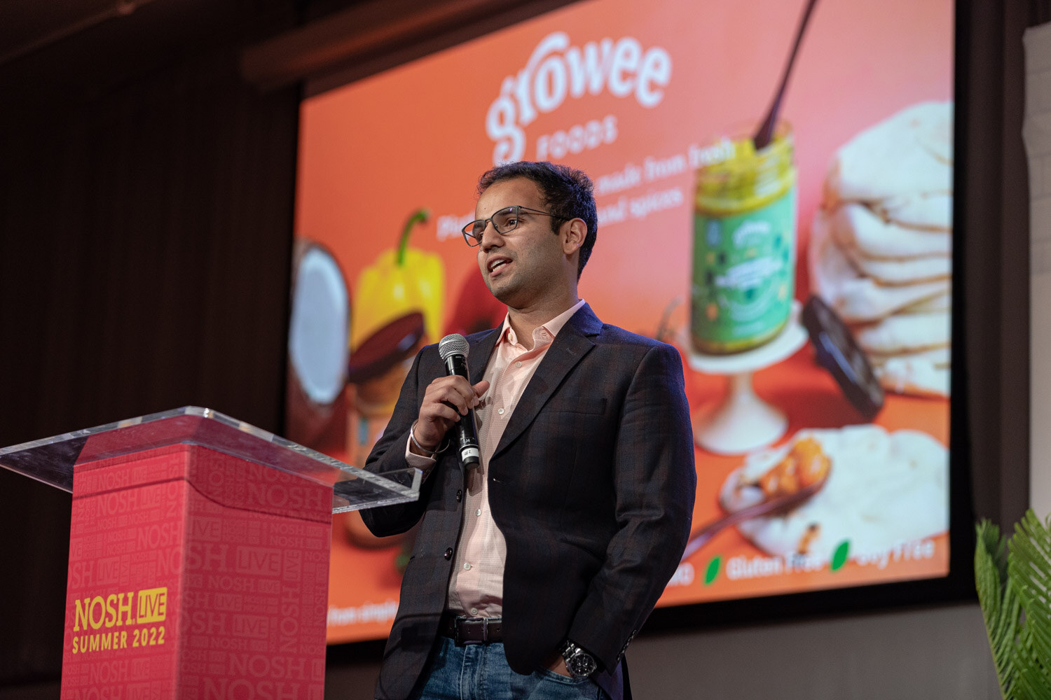 Man in glasses and a dark blazer with a mic at a red podium in front of an orange Growee Foods slide.