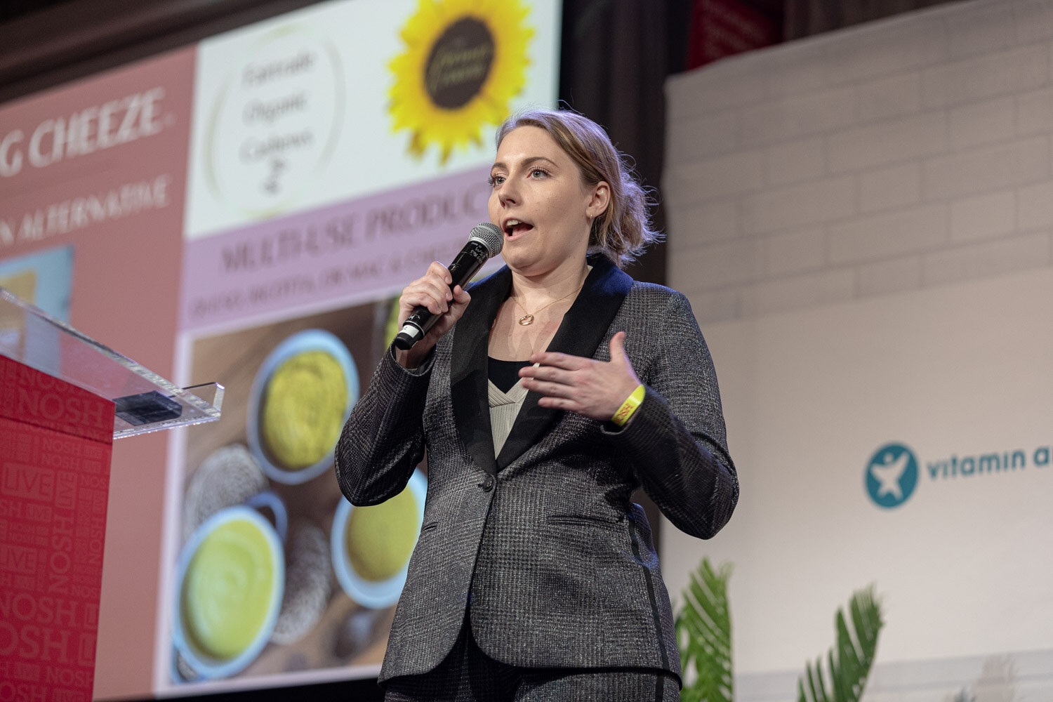 Woman in a dark gray blazer speaking into a mic beside a slide reading Everything Cheeze, cashew parmesan alternative.