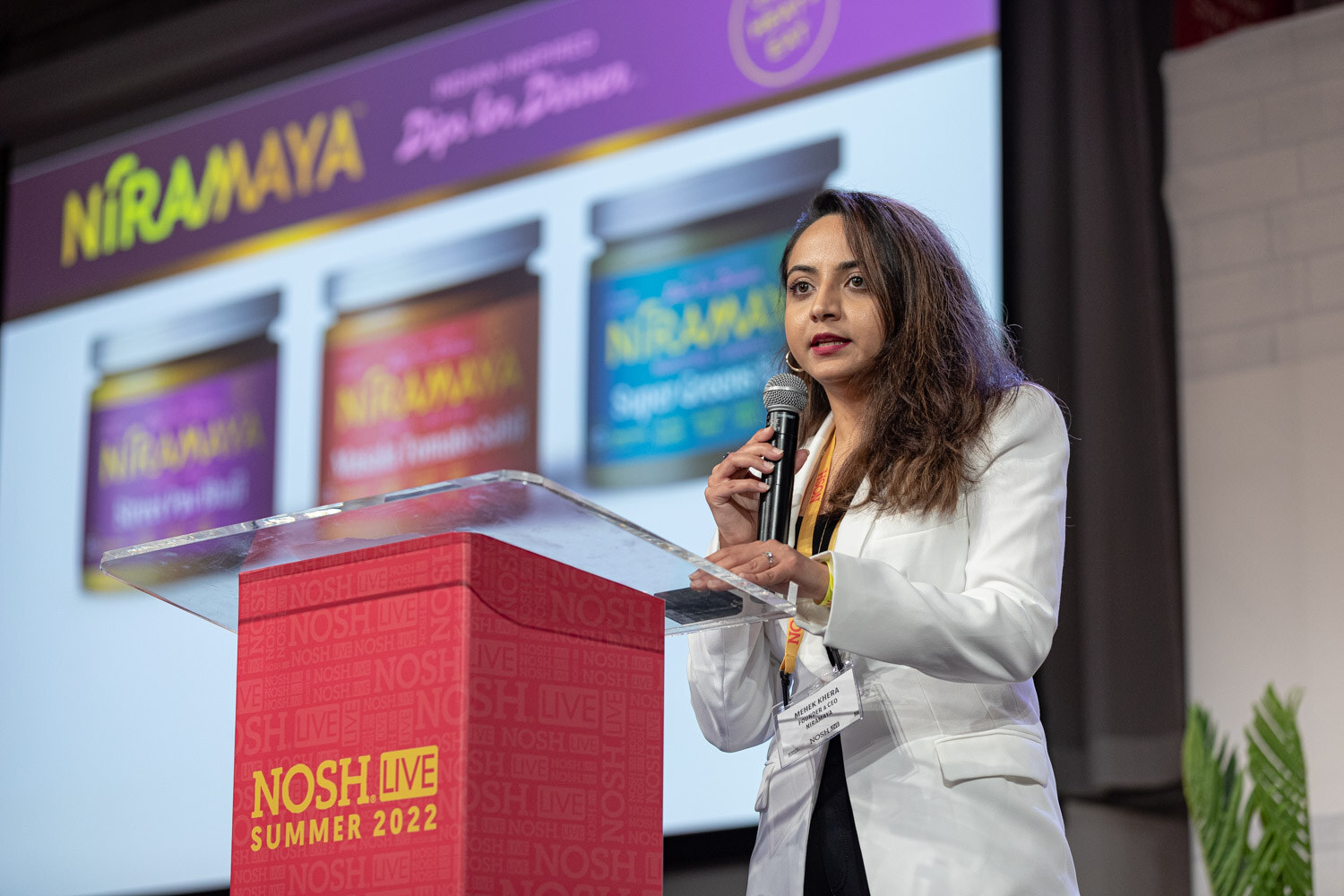 Woman in a white blazer holding a mic at a red podium in front of a slide showing three Niramaya jars.
