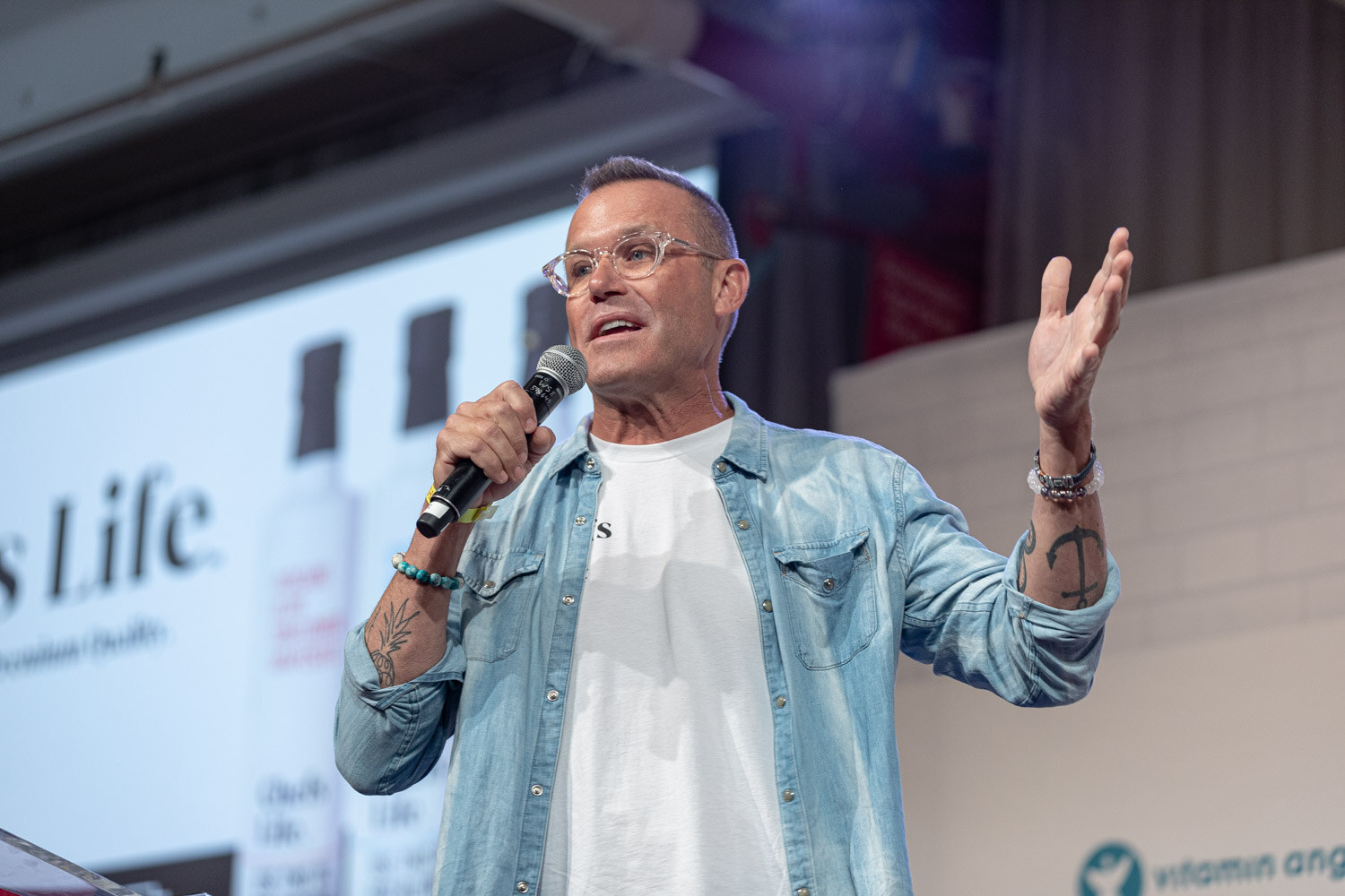 Man in glasses, a denim jacket and a white tee with a mic and one hand raised, a Chefs Life slide behind him.