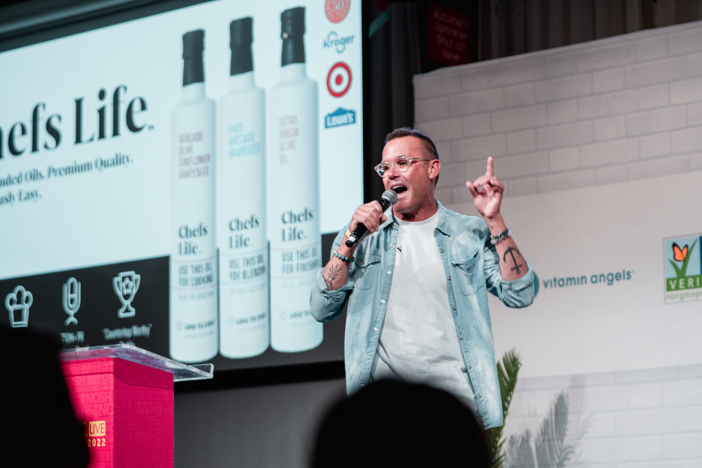 Man in a denim jacket holding a mic with one finger raised in front of a slide of Chefs Life oil bottles.