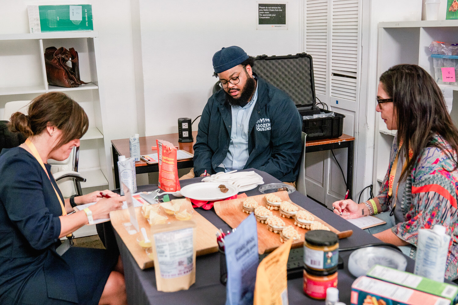 Three people seated around a table of tasting cups and snack packages, two writing and a man in an A Dozen Cousins jacket reading a package.