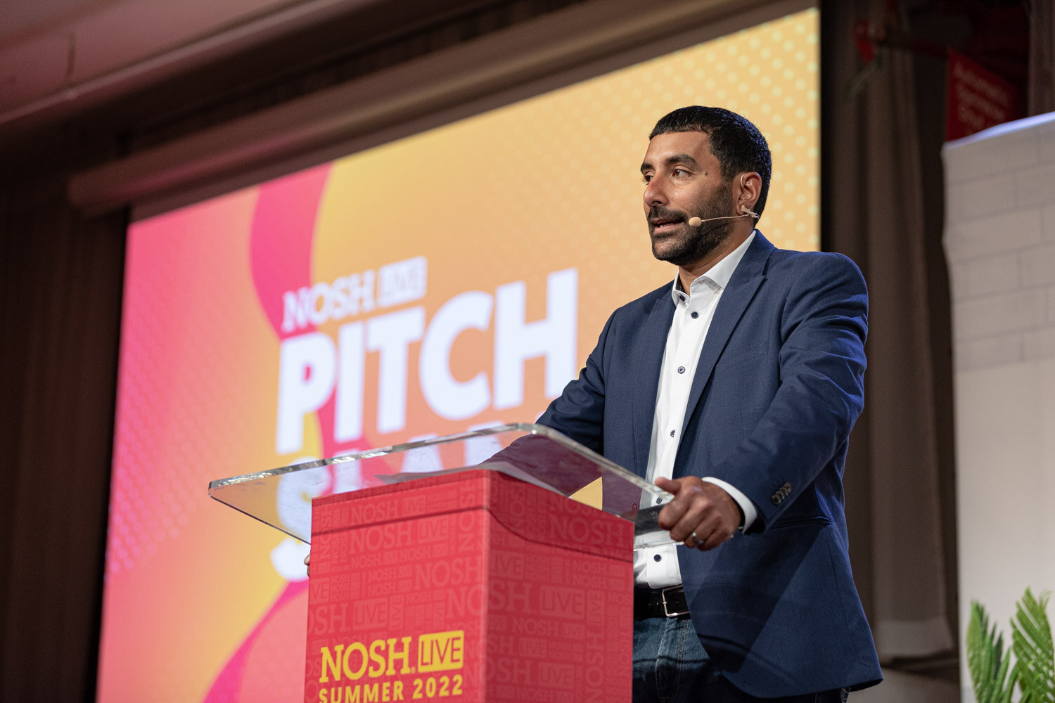 Man in a navy blazer and open-collar shirt speaking at a red podium in front of a Pitch Slam screen.