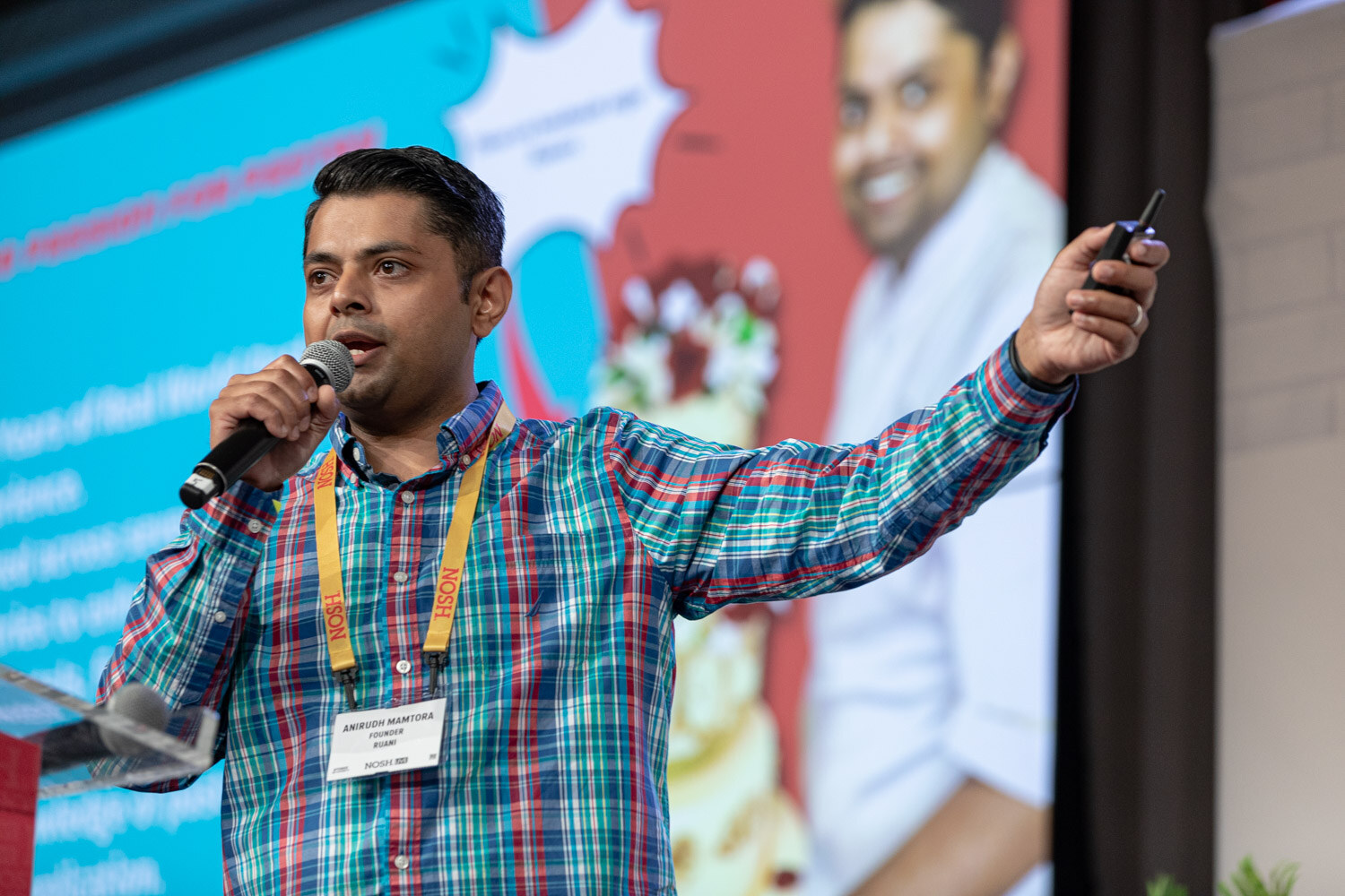 Man in a teal plaid shirt and yellow lanyard holding a mic and clicker, one arm stretched toward a slide behind him.