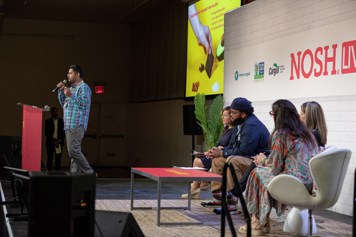 Wide stage view with a man in a plaid shirt speaking at the left and four judges seated in white chairs at the right.