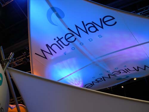 Large white fabric sails lit in blue and purple, printed with the WhiteWave Foods logo, hang above a trade-show booth with truss lighting in the dark ceiling.