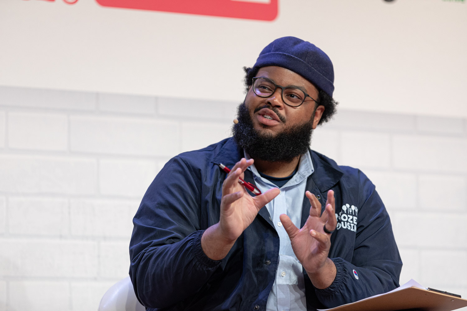 Bearded man in glasses, a navy beanie and a navy jacket gesturing with both hands while holding a clipboard.