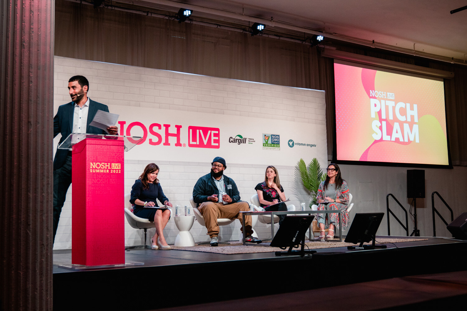 Wide stage with a man at a red podium, four seated judges, a Nosh Live backdrop and a Pitch Slam screen.