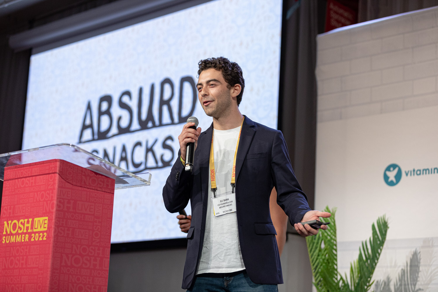 Man in a navy blazer and white tee with a mic beside a podium and a screen reading Absurd Snacks.