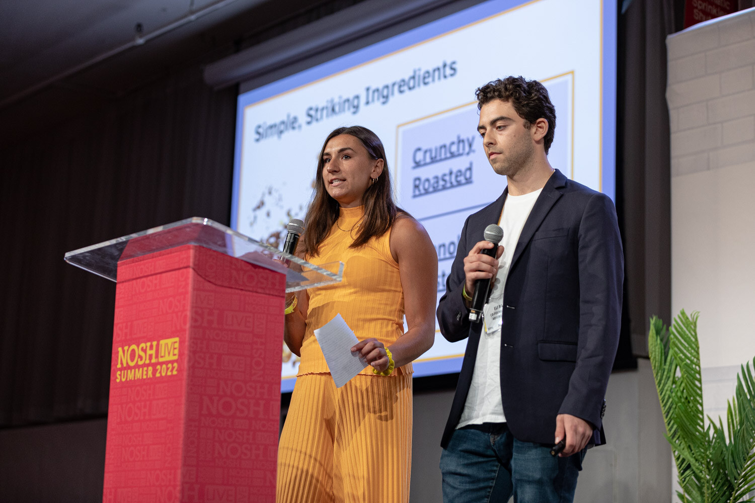 Woman in a yellow jumpsuit and man in a navy blazer holding mics beside a podium and a slide titled Simple, Striking Ingredients.
