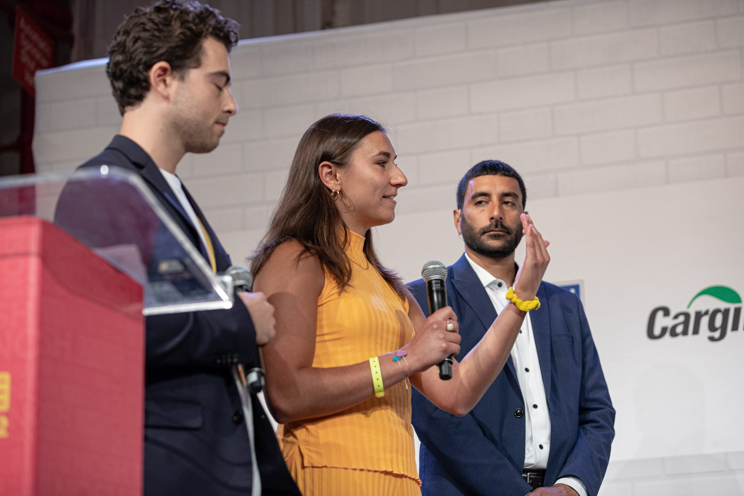 Woman in an orange top holding a mic with a man in a navy blazer and a bearded man beside her at a podium.
