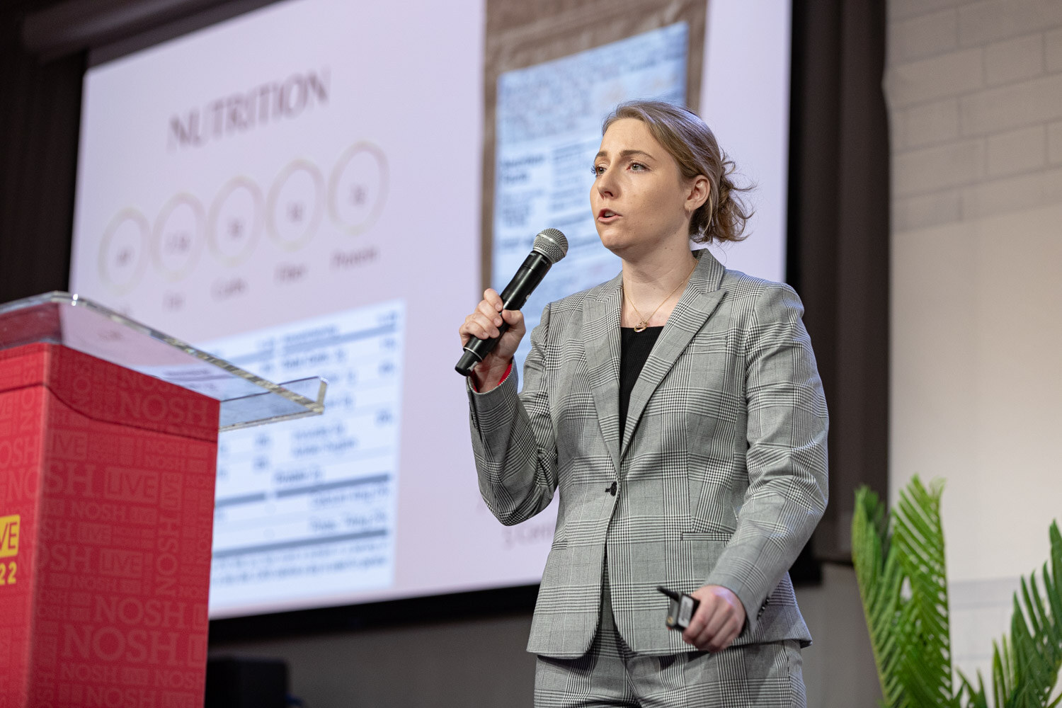 Woman in a gray plaid suit holding a mic and clicker in front of a screen showing a nutrition label.