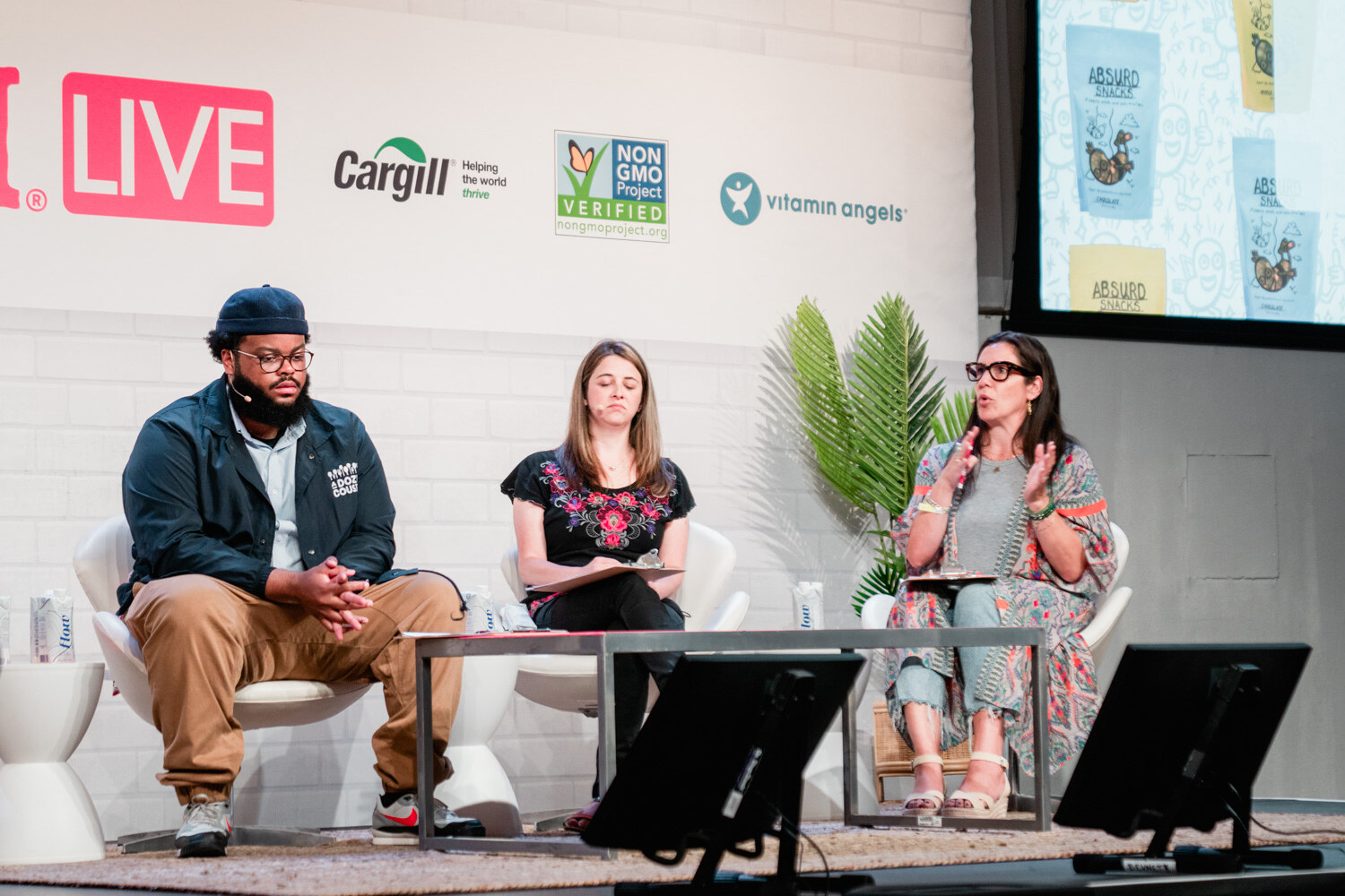 Three seated judges, a man in a navy jacket and two women, in front of a Nosh Live wall with palm plants.