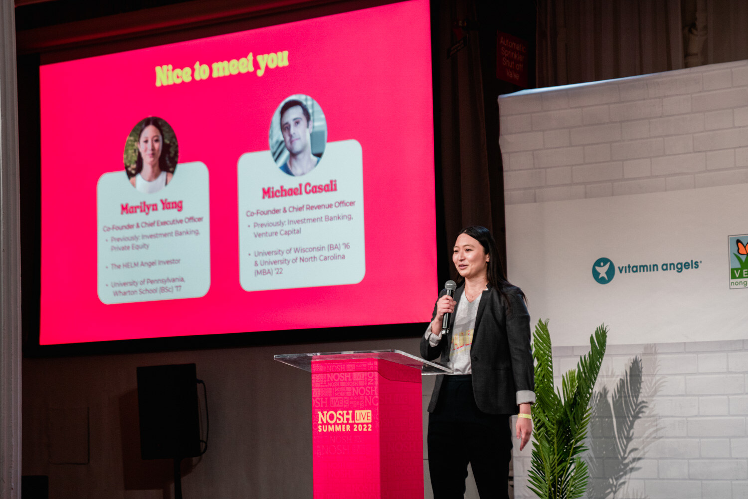 Woman in a dark blazer with a mic beside a red podium and a pink slide titled Nice to meet you.