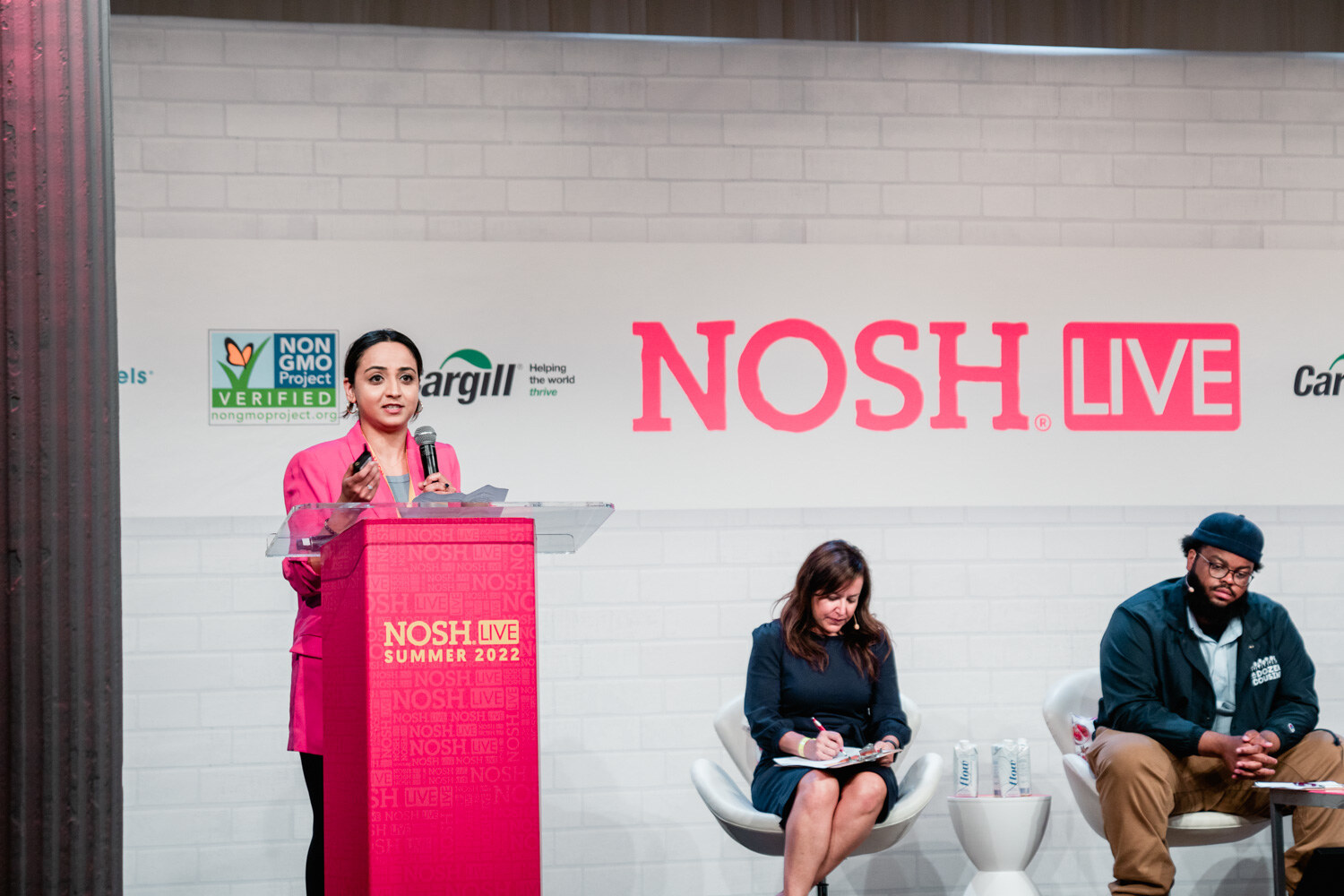 Woman in a pink blazer speaking at a red podium with two judges seated nearby under the Nosh Live logo.