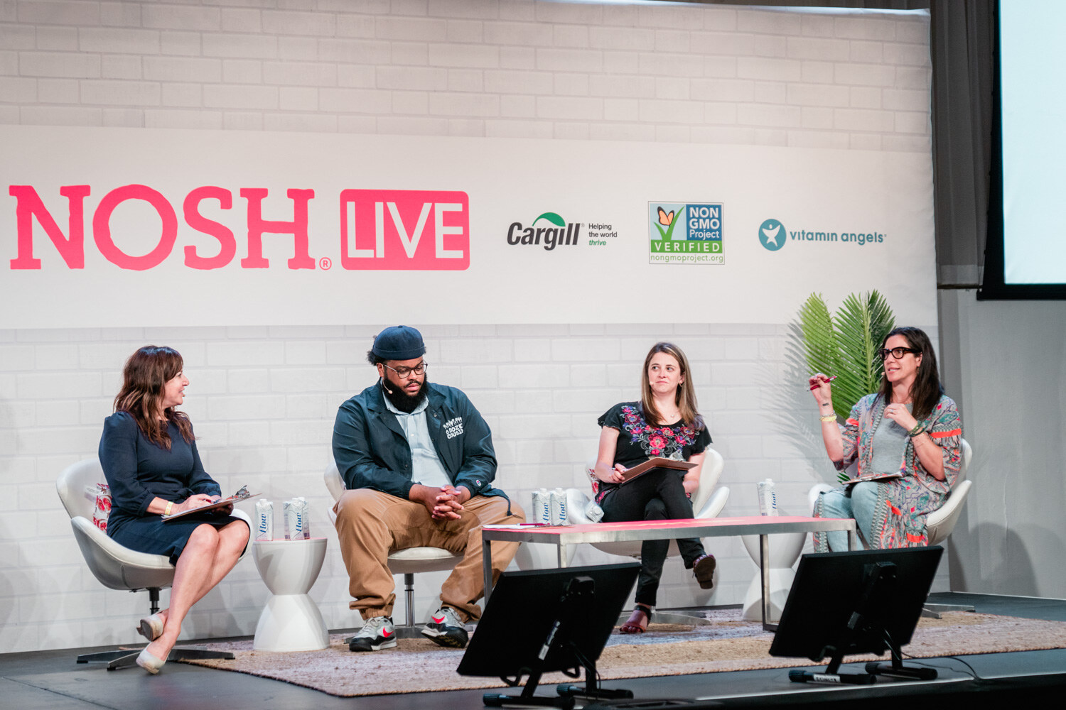Four seated judges in white chairs in front of a Nosh Live wall, one with a notebook on her lap.