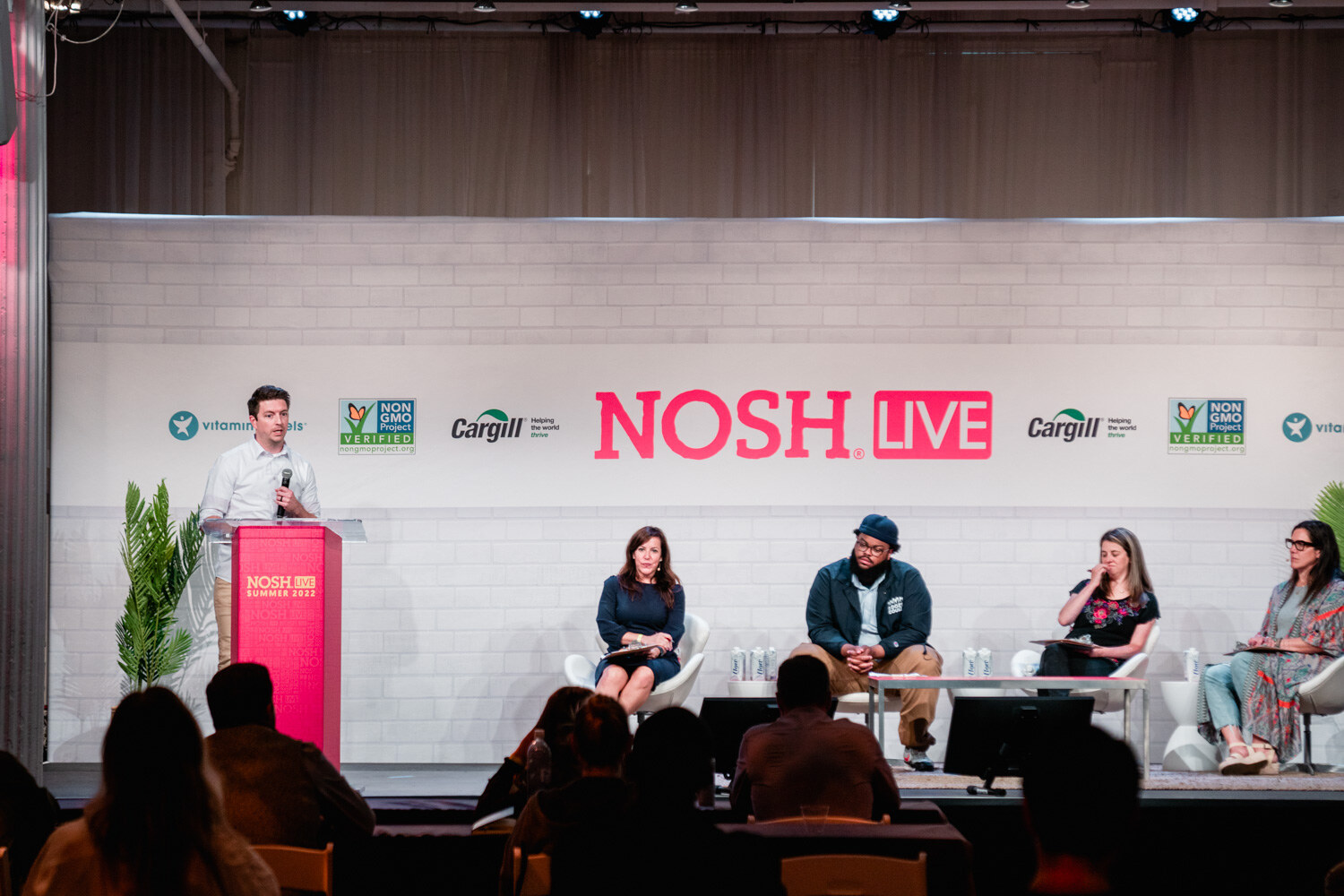 Wide stage with a presenter at the podium, four judges seated and audience heads in the foreground.