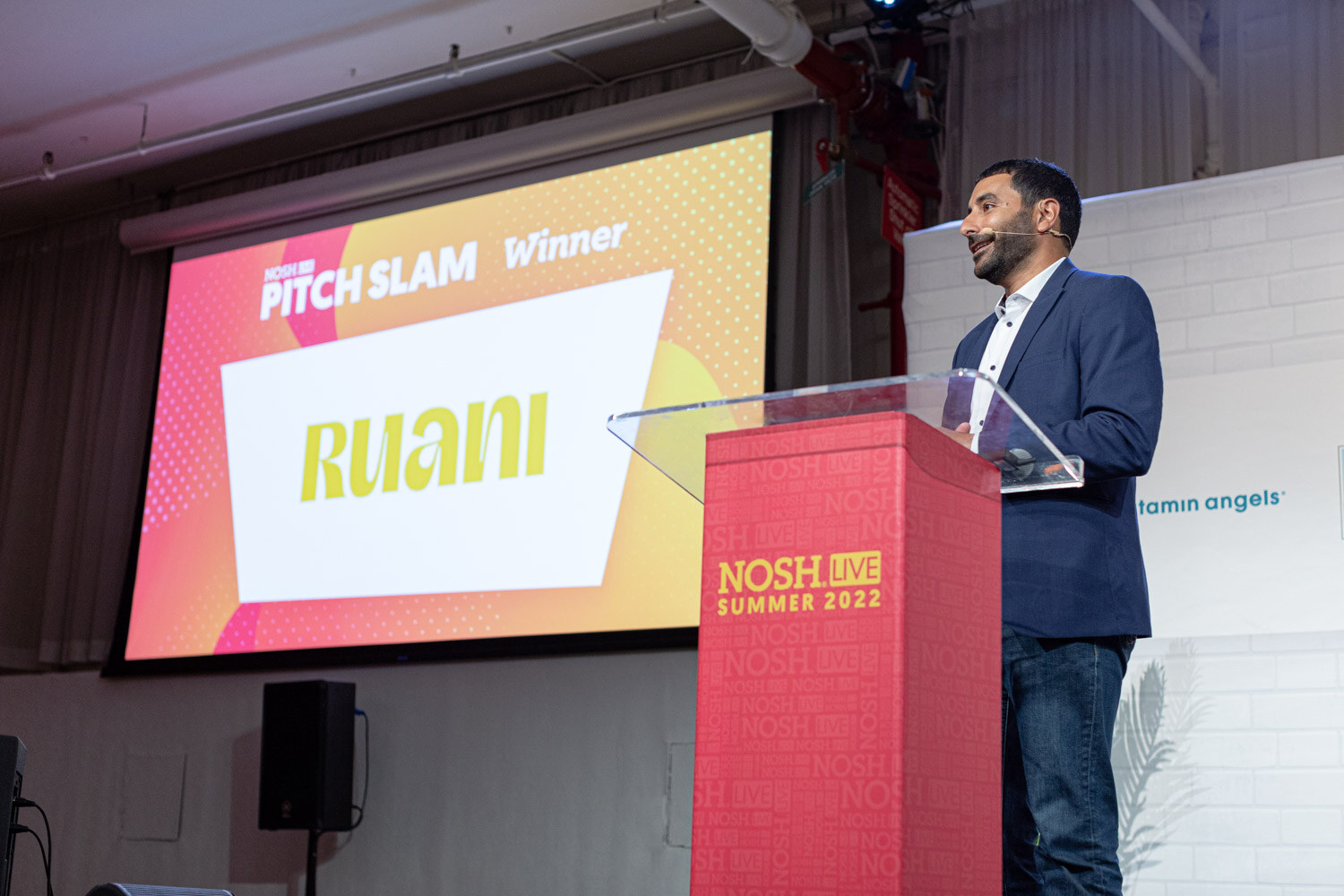 Man in a navy blazer at a red podium looking at a screen that reads Pitch Slam Winner Ruani.