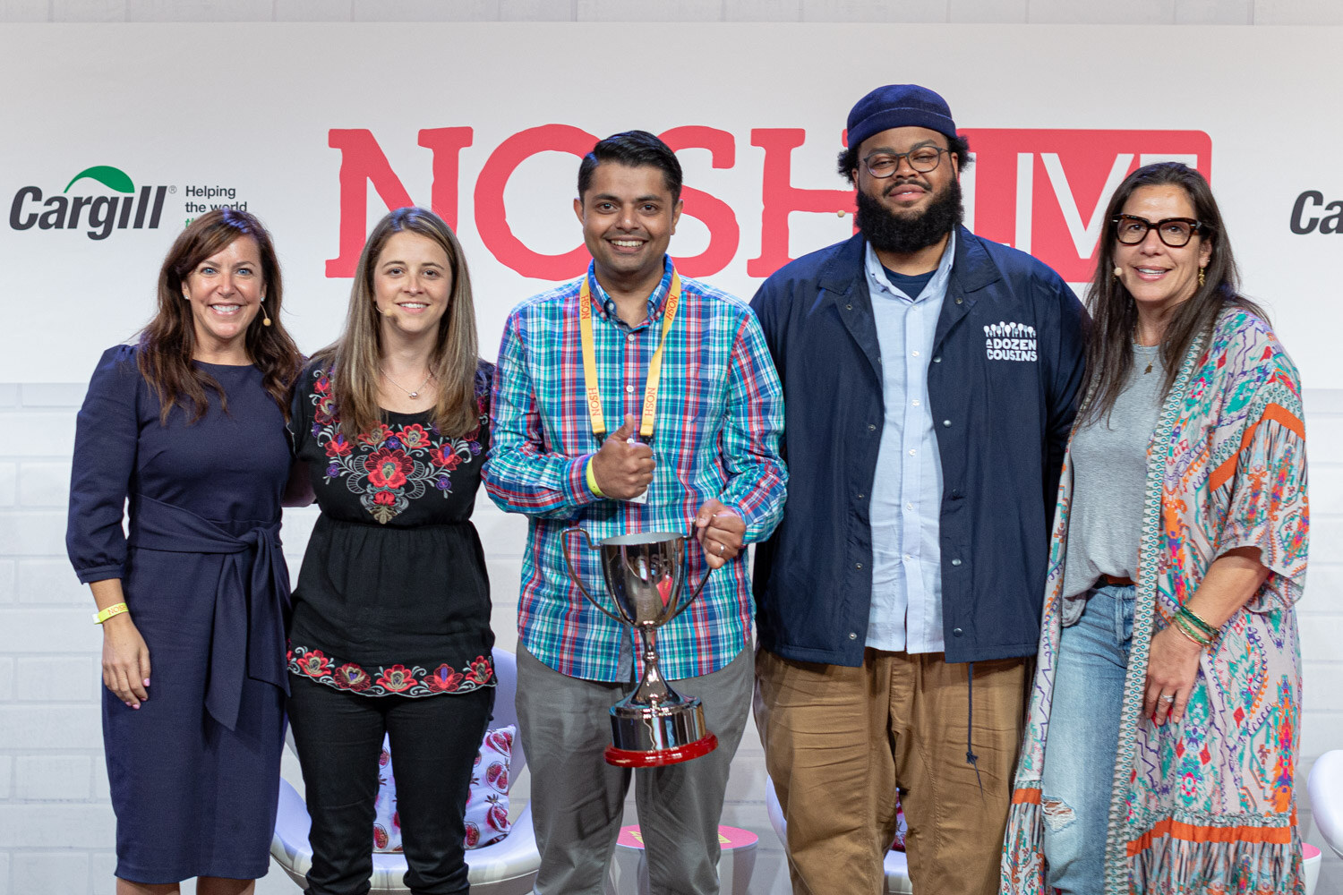 Five people smiling in front of a Nosh Live backdrop, one in the middle holding a silver trophy.