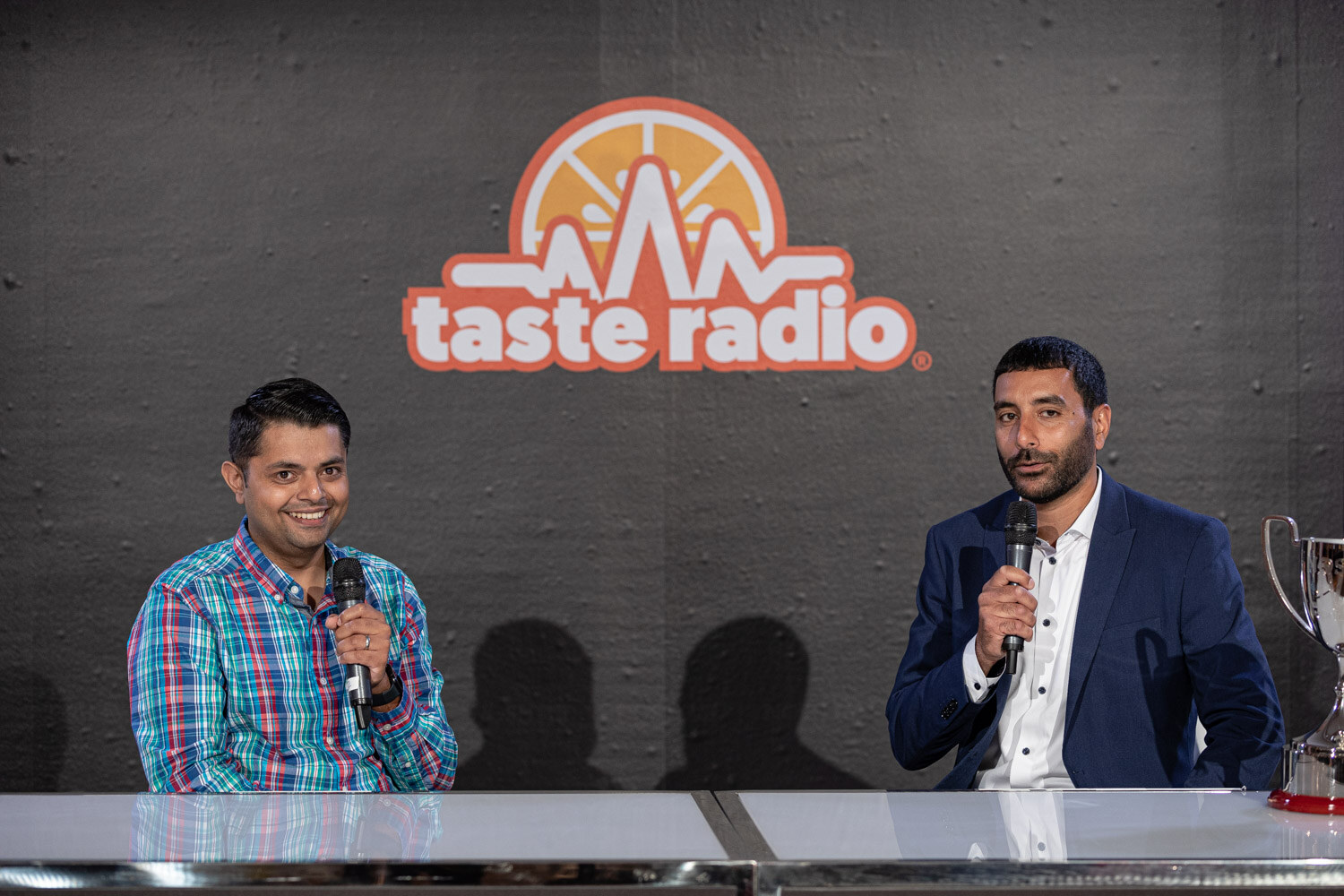Man in a plaid shirt and man in a navy blazer seated at a desk holding mics in front of the Taste Radio logo, a trophy at the right.