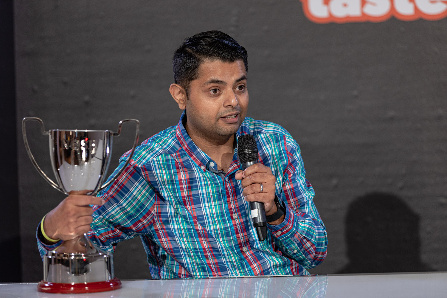 Man in a teal plaid shirt speaking into a mic with one hand on a large silver trophy on the desk.