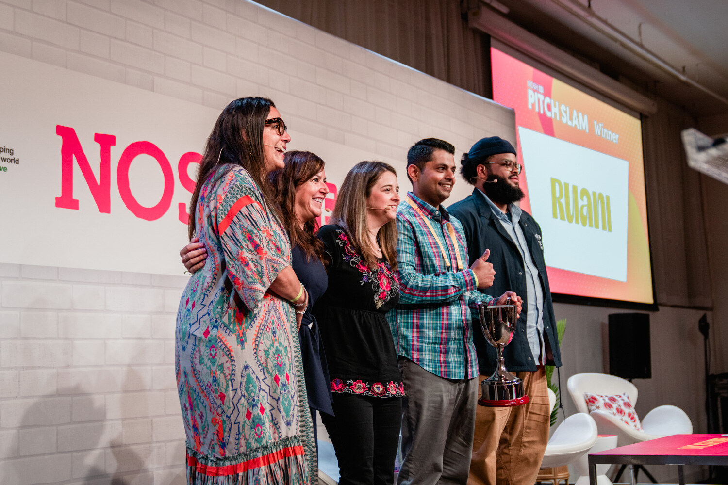 Five people standing together on stage, one holding a silver trophy, with a Pitch Slam Winner Ruani slide behind them.