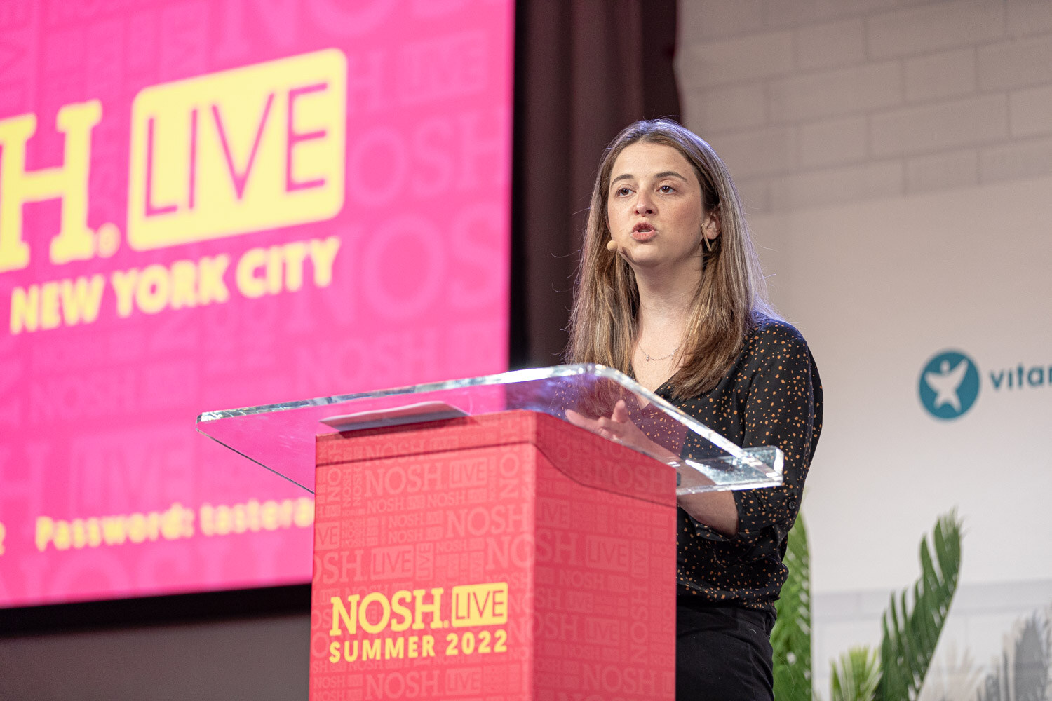 Woman with a mic at a clear podium in front of a pink screen reading Nosh Live New York City.