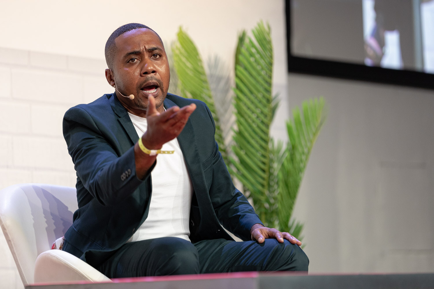 Man in a dark blazer and white tee seated on a white chair with one hand stretched out, palm up, beside a large palm plant.